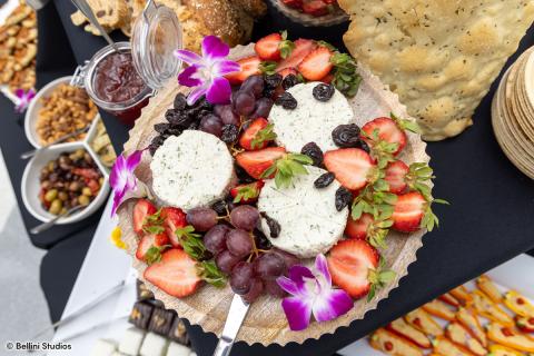 Close up of a cheese spread plate at the Presidential Farewell Reception at University at Buffalo's Center for the Arts, including grapes, strawberries, and raisins. Little purple flowers decorate the plate as well. There are bowls of olives, nuts and jam in the background. Photo by Bellini Studios.