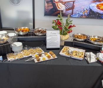 A vegetarian and vegan option table decorated for autumn with flowers and fall leaves. An assortment of different food items including chocolate chip cookies, mini muffins, tempura bites, and flatbread are on display ready for serving.