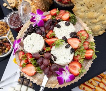 Close up of a cheese spread plate at the Presidential Farewell Reception at University at Buffalo's Center for the Arts, including grapes, strawberries, and raisins. Little purple flowers decorate the plate as well. There are bowls of olives, nuts and jam in the background. Photo by Bellini Studios.